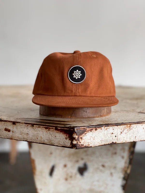 The Ballad of the Bird Dog Mescalito Field Hat rests on a worn, white metal table at Mescalito Coffee—a brown cap with a black circular patch and white geometric design.