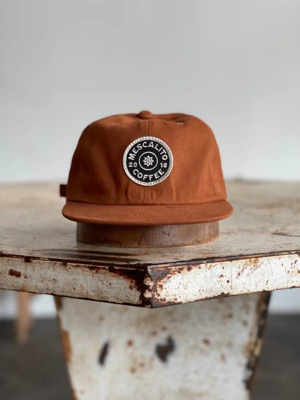 The Mescalito Field Hat by Ballad of the Bird Dog features a brown cotton design and a circular “Mescalito Coffee” patch—an ideal addition to any coffee shop hat collection.