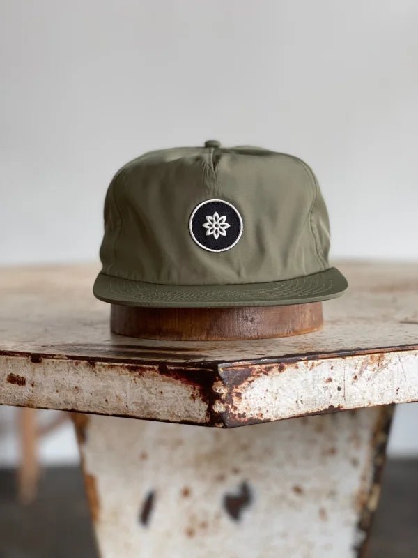 The Mescalito Nylon Field Hat by Ballad of the Bird Dog, featuring a black and white flower emblem, rests stylishly on a distressed wooden table—a nod to Mescalito Coffee’s vibe.