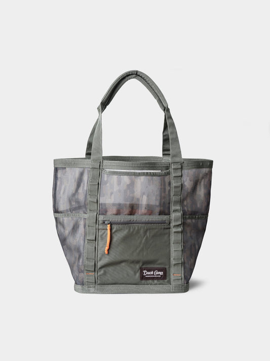 The Duck Camp Mesh Gear Tote is a green, water-resistant bag with camouflage mesh panels, dual handles, an exterior zip pocket featuring an orange pull, and a “Duck Camp” label on the front.