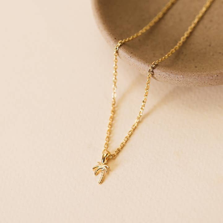 The JaxKelly Mini Palm Tree Pendant Necklace, 18k gold plated and hypoallergenic, features a small pendant and is shown on a beige surface beside a round brown dish.