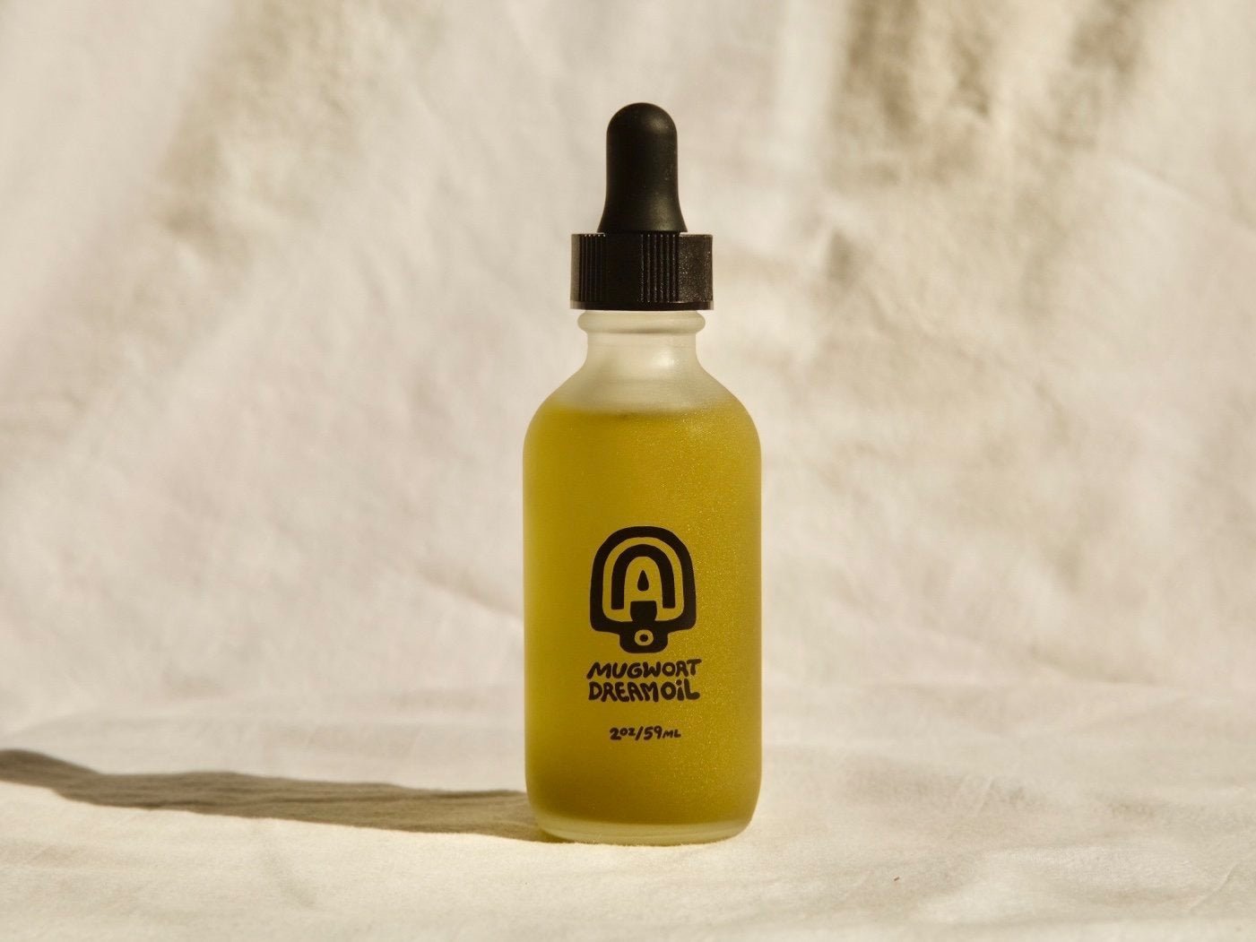 A clear glass dropper bottle filled with yellow-green liquid labeled "Mugwort Dream Oil | Mother Mountain Herbals" rests on a white fabric surface—ideal as a relaxing massage oil or botanical body oil from Mother Mountain Herbals.