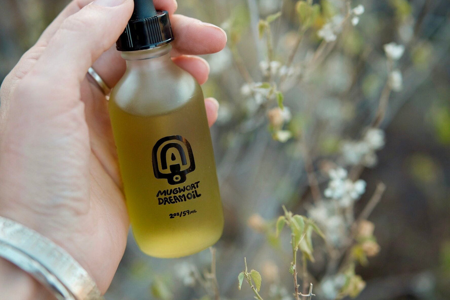 A hand holds a glass dropper bottle of Mugwort Dream Oil by Mother Mountain Herbals, a botanical body oil, outdoors near blurred plants.