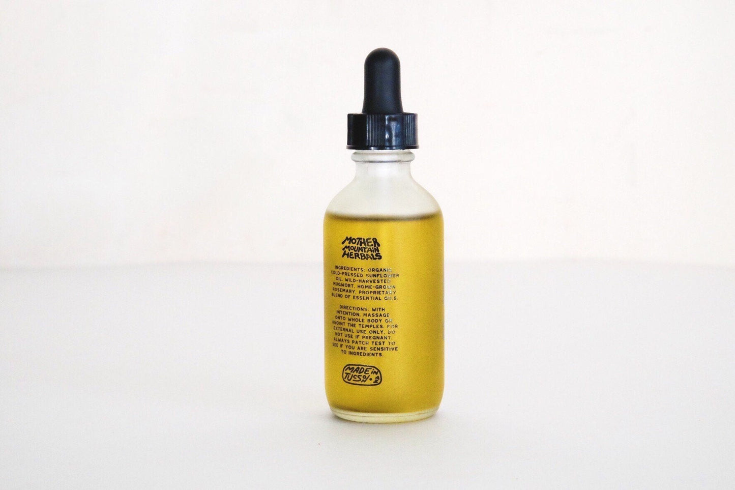 A glass dropper bottle filled with yellow liquid, labeled "Mugwort Dream Oil | Mother Mountain Herbals," stands upright on a plain white surface—ideal as a botanical body oil.