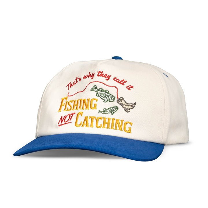 The Not Catching Hat by Sendero Provisions Co. is a white and blue unstructured 5-panel fishing hat featuring embroidered twill text, “That's why they call it Fishing NOT Catching,” and two fish graphics.