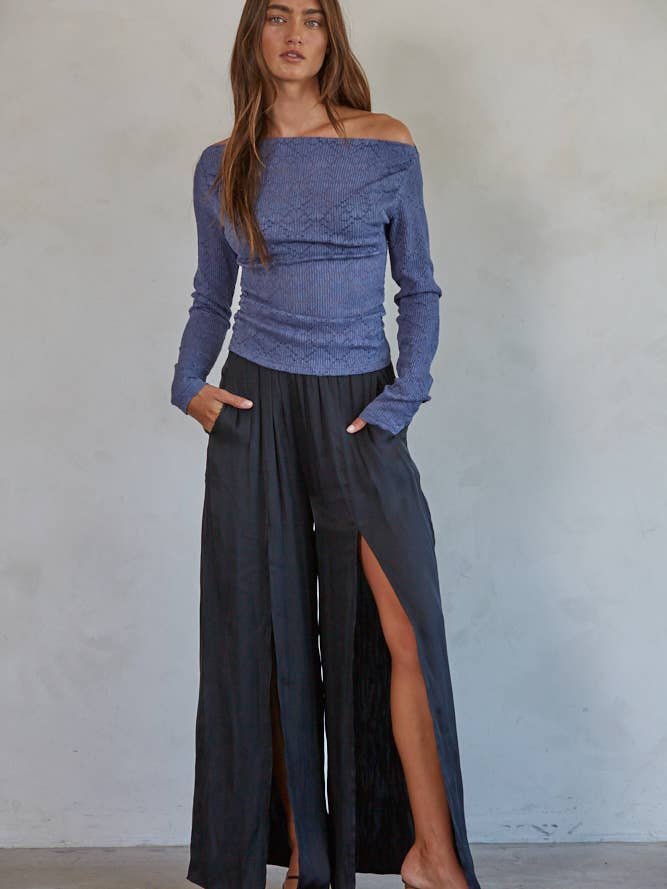 A woman stands against a plain wall wearing the By Together Off The Shoulder Top, paired with black wide-leg pants featuring high slits and her hands tucked casually in her pockets.