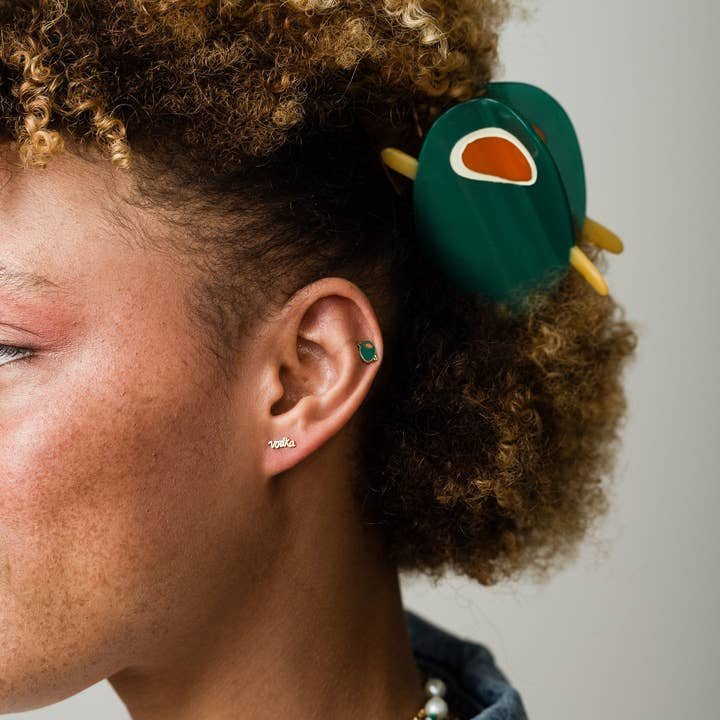 Close-up of a person with curly hair wearing the Olive Claw Clip by Larissa Loden, featuring a green and orange patterned cellulose acetate design, along with small stud earrings—one reads "cool.
