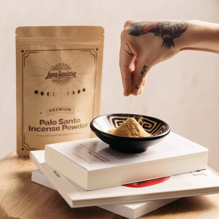 A hand sprinkles Luna Sundara Palo Santo Incense Powder into a bowl, releasing its aromatic scent. The product packet and stacked books are displayed on a wooden surface.