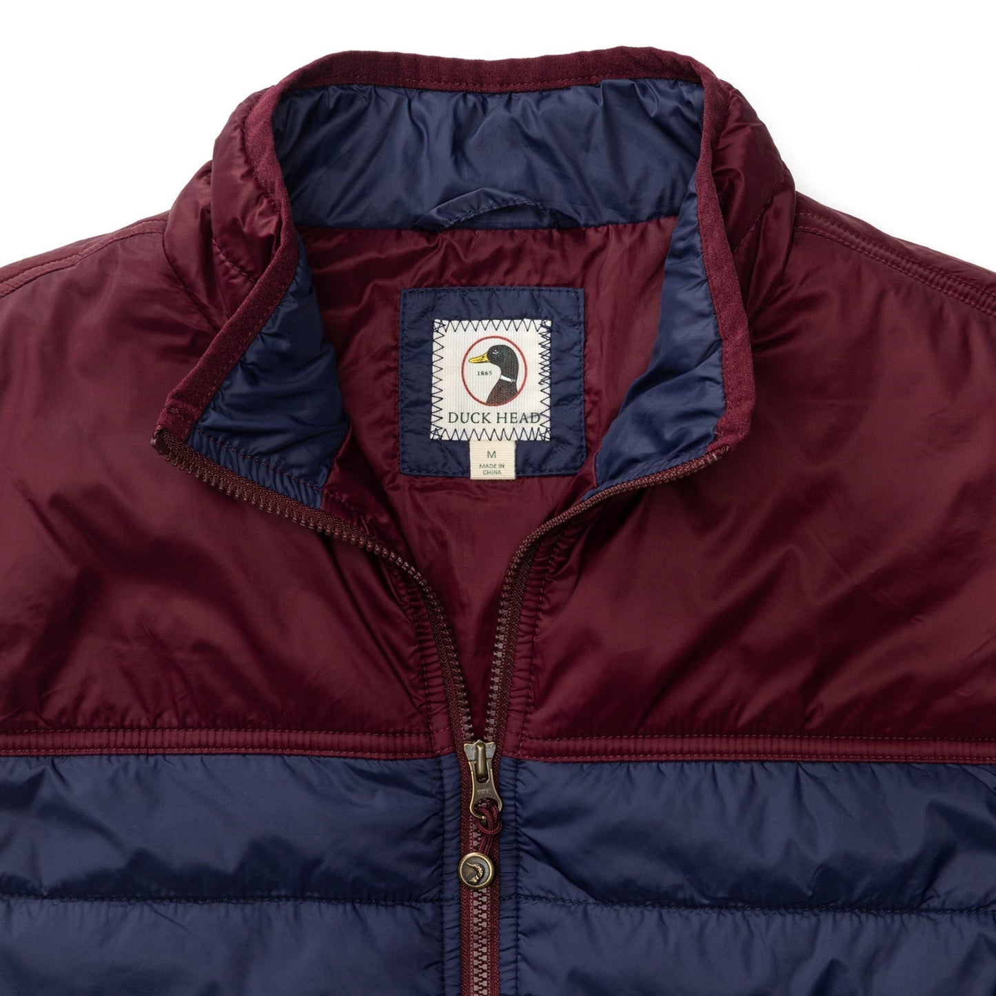 A close-up of the Duck Head Ridgeland Performance Quilted Vest in maroon and navy with PrimaLoft insulation, a zipper, and the "Duck Head" label inside the collar.