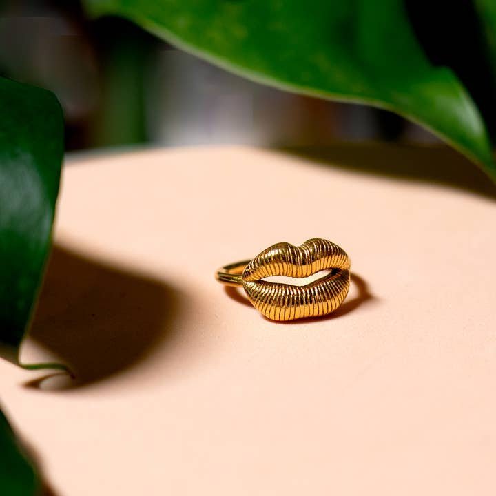 The Ring | Gold Lips by Peter and June features a textured lip design in ethically sourced, 18k gold plated material. This adjustable piece rests on a pale surface with green leaves, adding conscious elegance to your look.