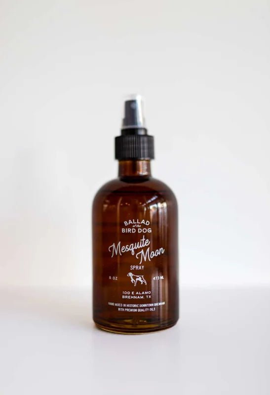 A brown glass bottle labeled "Room & Linen Spray | Mesquite Moon | Ballad of the Bird Dog" by Ballad of the Bird Dog, made with essential oils, sits on a white surface—ideal for those seeking a unique room or linen spray.