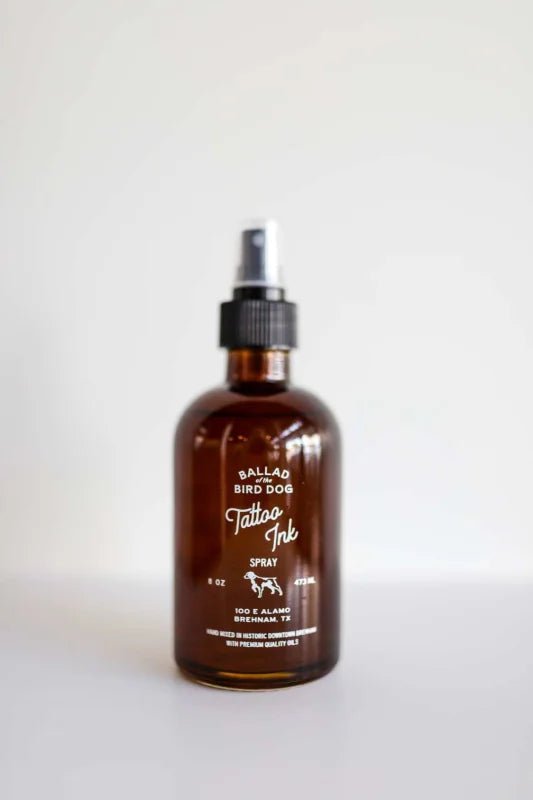 A brown glass bottle labeled “Room & Linen Spray | Tattoo Ink” by Ballad of the Bird Dog, featuring a black pump top—perfect for freshening rooms and linens—displayed against a plain white background.
