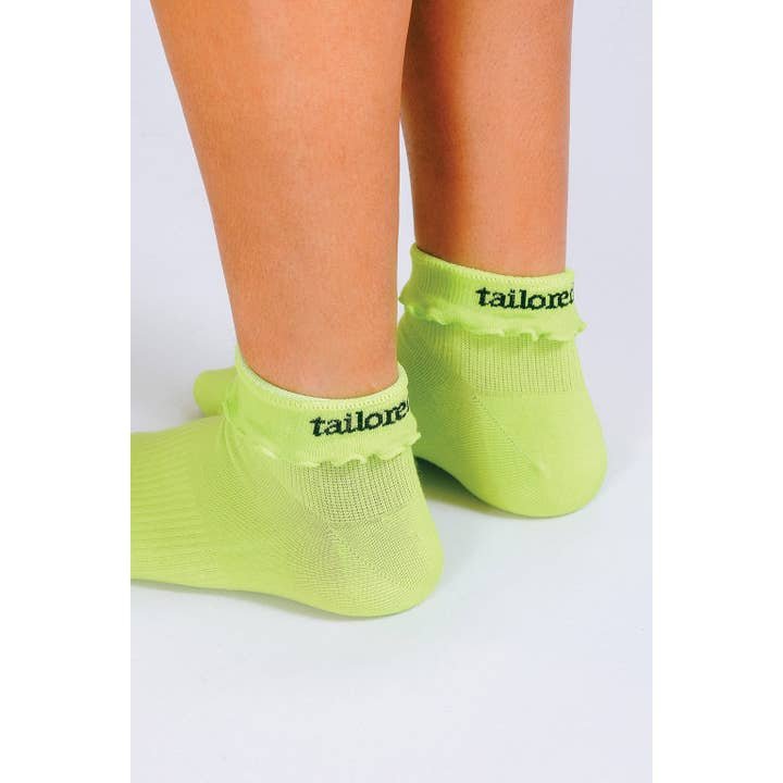 A person wearing Tailored Union's Ruffle Ankle Sock—lightweight neon green socks with reinforced heel and toe, featuring "tailored" on the back.