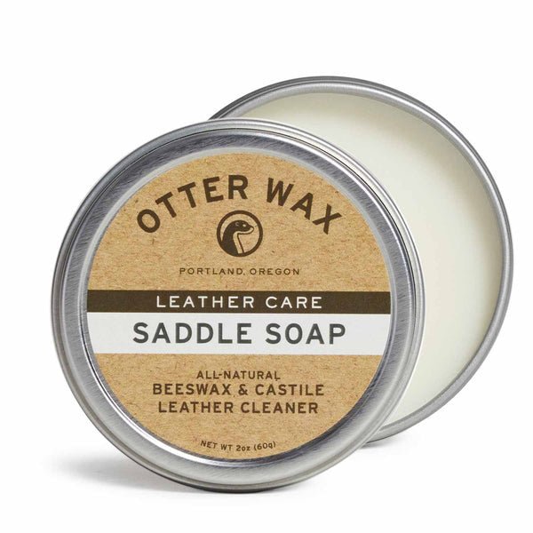 A round tin labeled "Saddle Soap | Otter Wax" from Otter Wax sits with its lid partially off, revealing the solid, all-natural leather cleaner inside.
