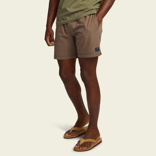 A person wearing an olive green shirt, Howler Brothers Salado Seersucker Shorts in brown, and tan flip-flops stands with one hand in their pocket against a plain background.