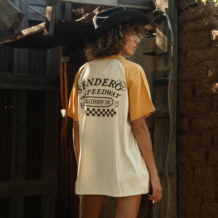 A person in a Sendero Provisions Co. Saloon Shirt, featuring a classic fit and "Fender Speedway Cowboy 500" graphic, stands outdoors near rustic wooden and stone structures.