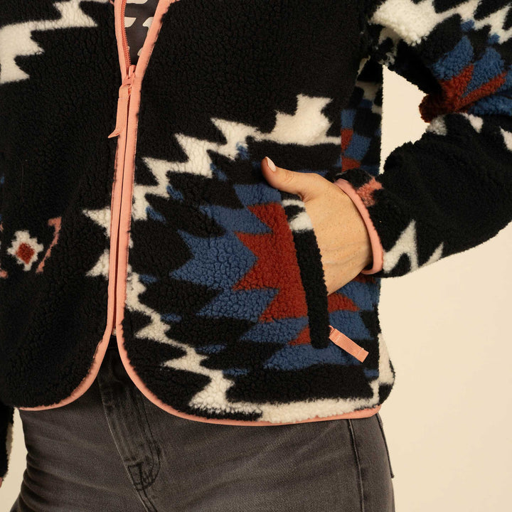 A person wearing the Sedona Fleece by Sendero Provisions Co.—a black recycled polyester jacket with a Southwest print, pink trim, front zipper, and side pocket—rests their hand in the pocket.