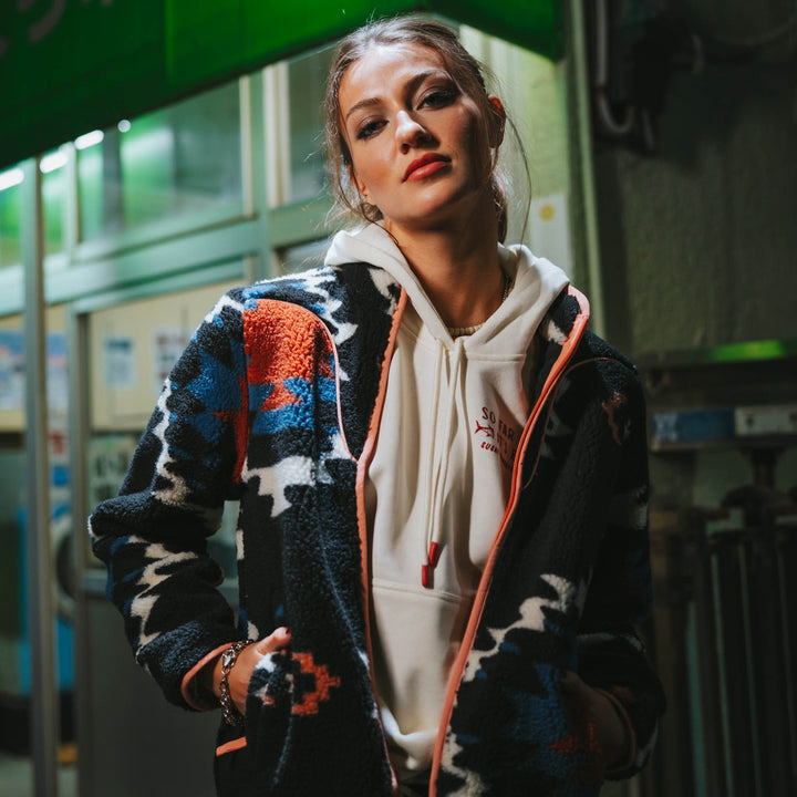 A woman in the Sendero Provisions Co. Sedona Fleece, made from recycled polyester, wears it over a white hoodie and stands indoors under fluorescent lights, looking directly at the camera.
