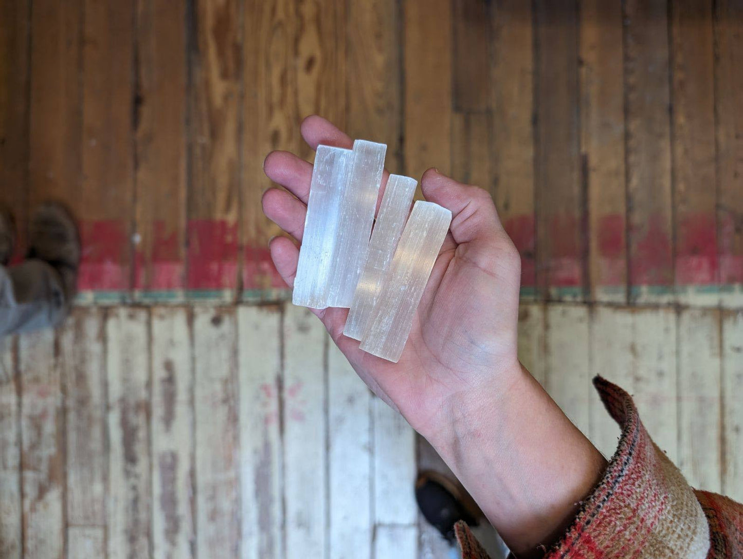 A hand holds three translucent Selenite Crystal Stacks from World Incense Store, ideal for crystal healing or chakra work, over a weathered wooden floor with red paint along the edge.