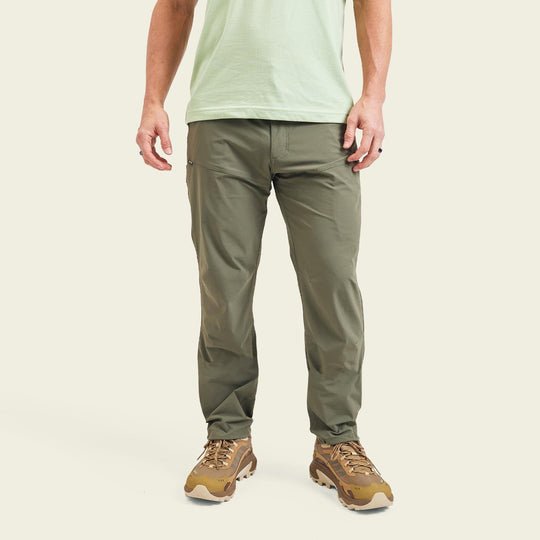 A person wearing a light green t-shirt, Howler Brothers Shoalwater Tech Pants in olive green, and brown sneakers stands against a plain light background.