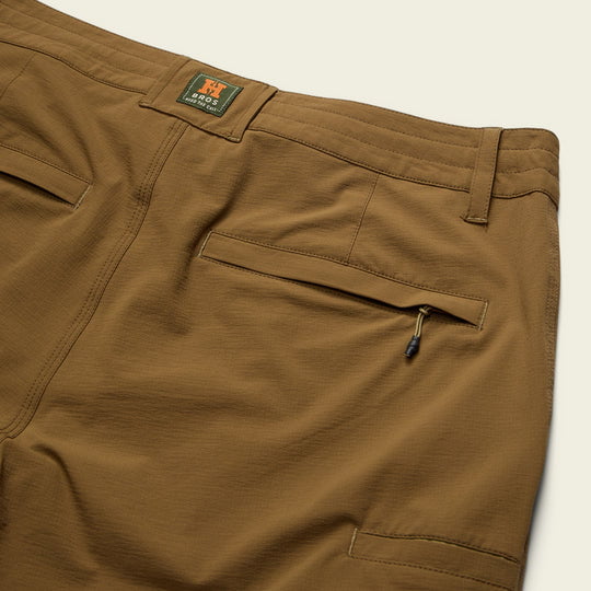 Close-up of the back of Shoalwater Tech Pants by Howler Brothers, showing two back pockets (one zippered), a belt loop, and a green Howler Brothers label above the waistband.