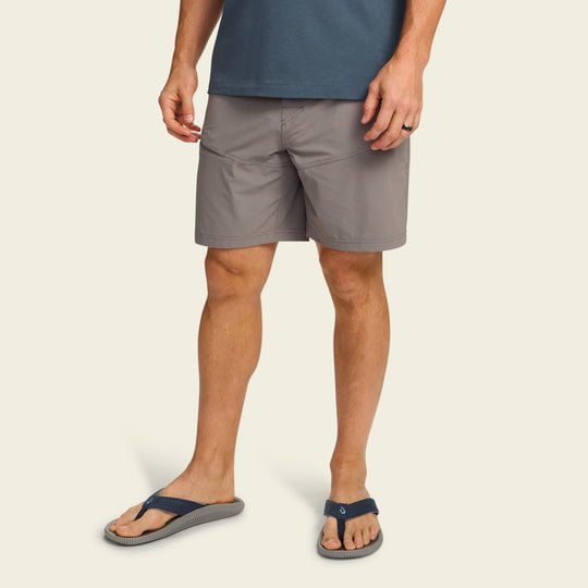 A man in a blue t-shirt, Howler Brothers Shoalwater Tech Shorts, and navy flip-flops stands against a plain light background.
