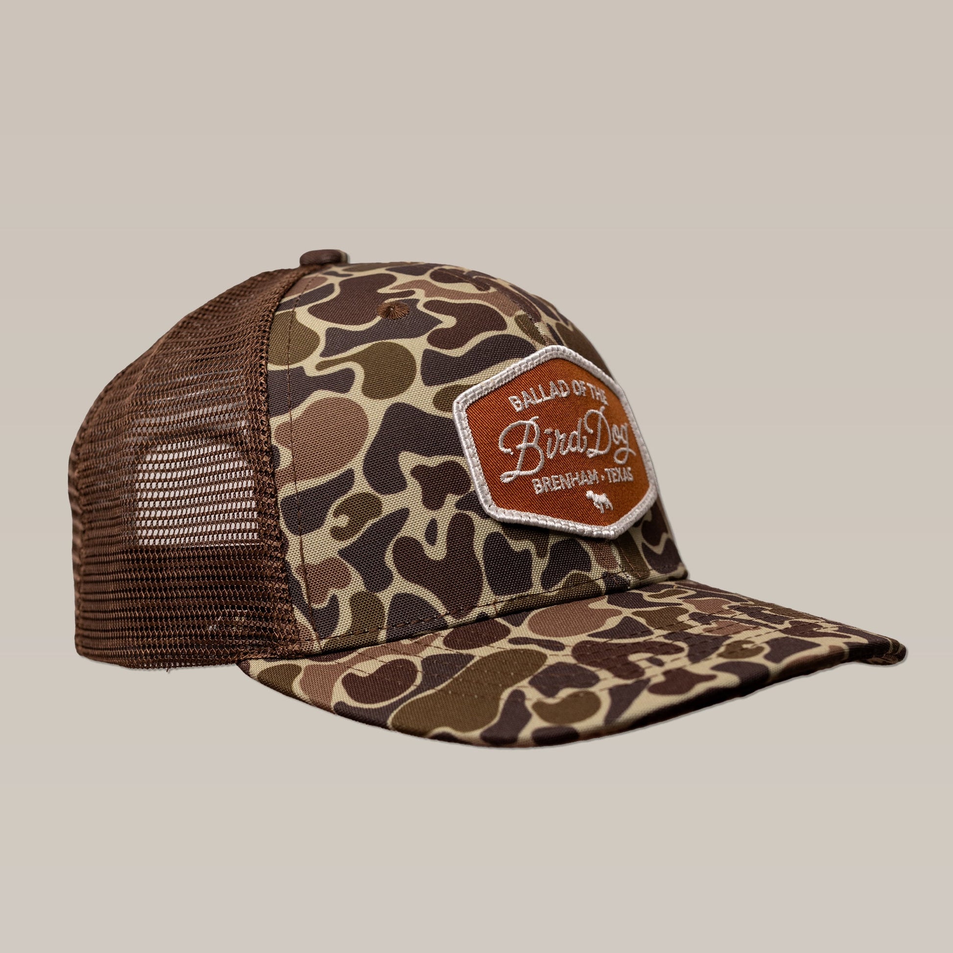 The Ballad of the Bird Dog Shop Hat | Badge is a brown and green camo trucker hat with mesh back, featuring a front patch that reads "Ballad of the Bird Dog, Brenham, Texas.