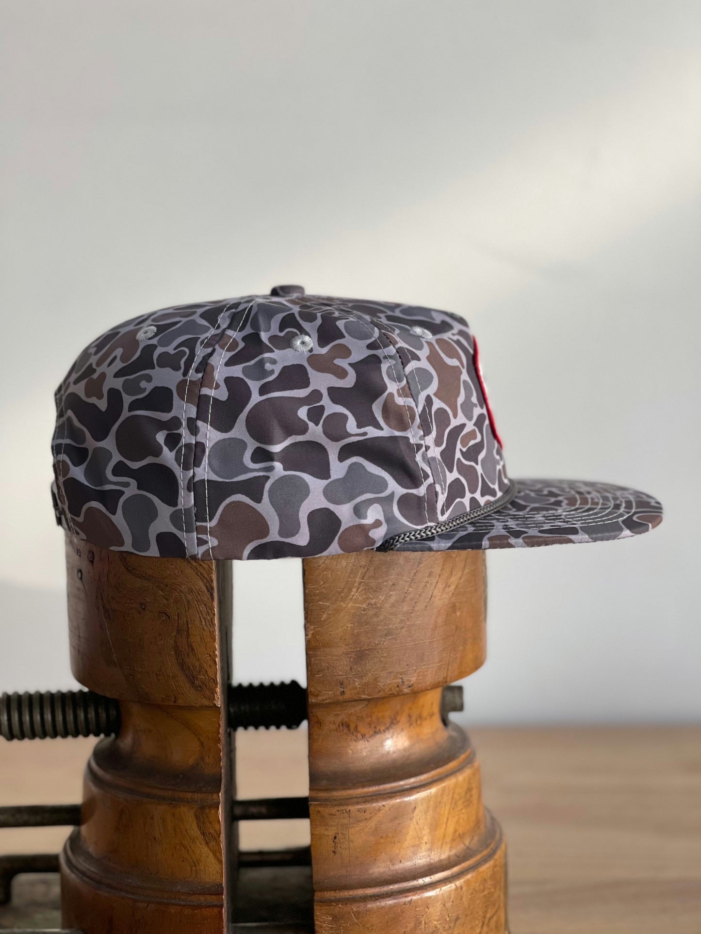 The Shop Hat | Camo Companion by Ballad of the Bird Dog in the Smoke Show Camo pattern sits on a vintage wooden hat stretcher—your ideal outdoor companion for any backdrop.