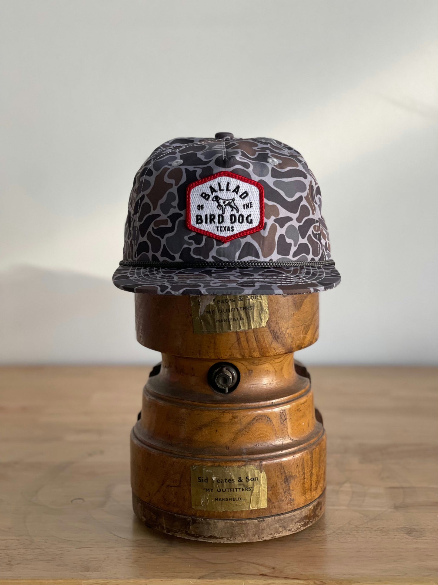 The Ballad of the Bird Dog Shop Hat | Camo Companion rests on a wooden hat block atop a rustic surface—the ideal outdoor accessory.