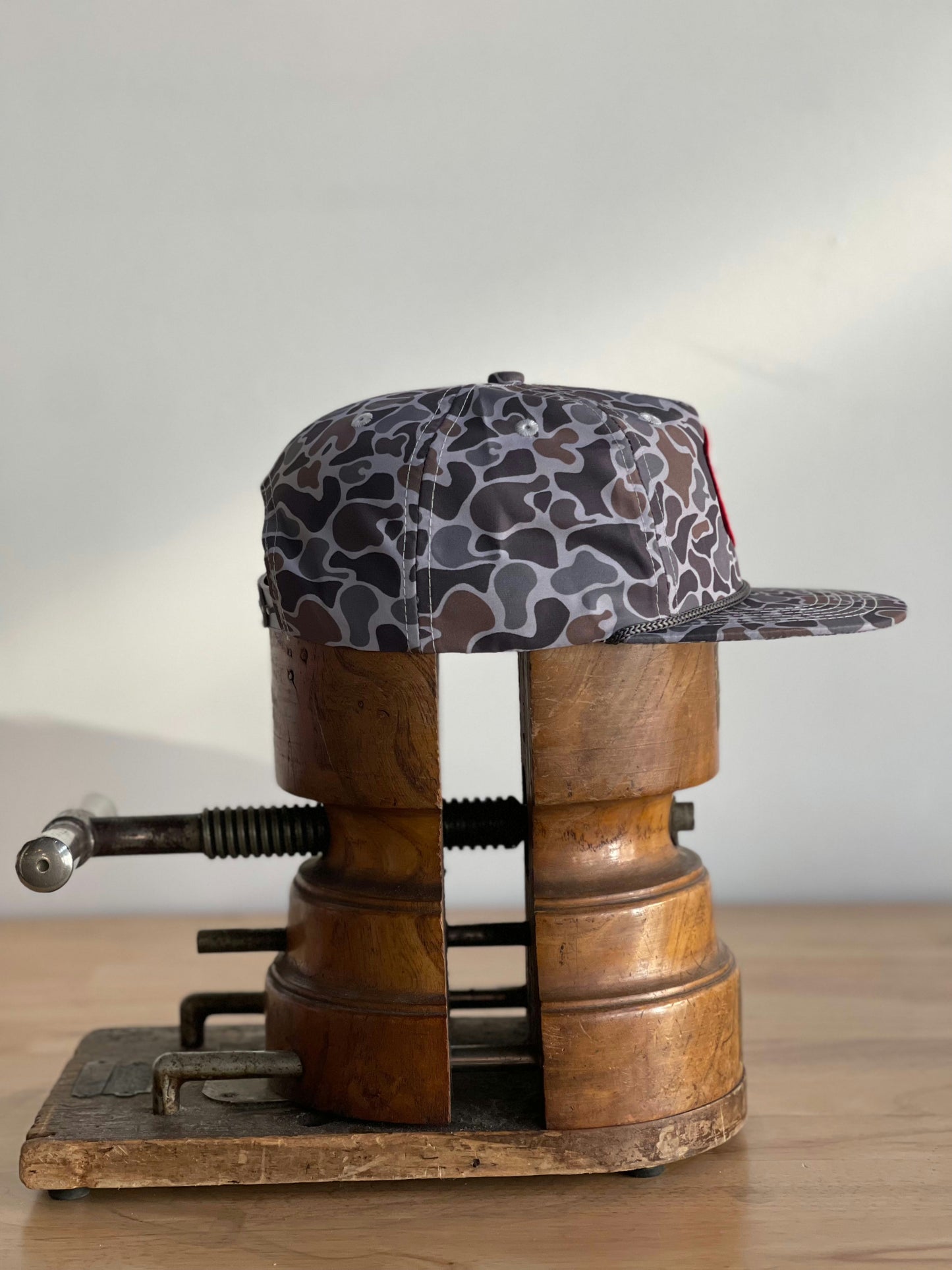 The Shop Hat | Camo Companion by Ballad of the Bird Dog stands upright in a vintage wooden clamp on a wood surface, showcasing its camouflage pattern—a perfect mix of rugged style and practicality against a plain background.