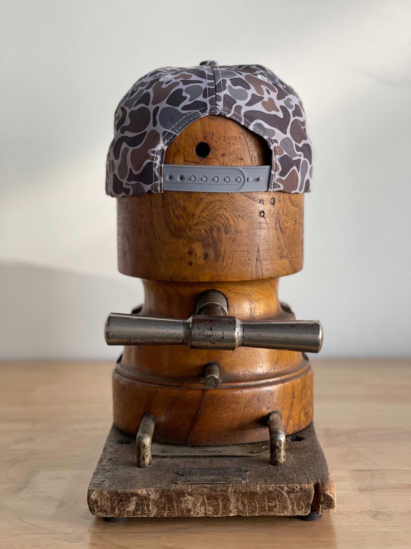 The Ballad of the Bird Dog Shop Hat | Camo Companion is shown from the back on a wooden hat block stand atop a wooden surface.