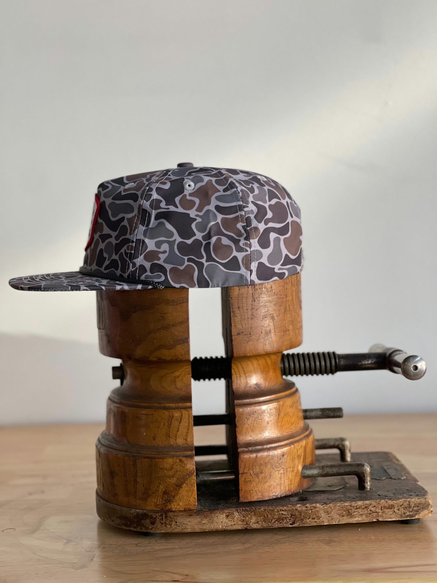 The Shop Hat | Camo Companion by Ballad of the Bird Dog is shown on a vintage wooden hat stretcher, atop a wood surface with a neutral backdrop—an ideal choice for outdoor enthusiasts seeking classic style.