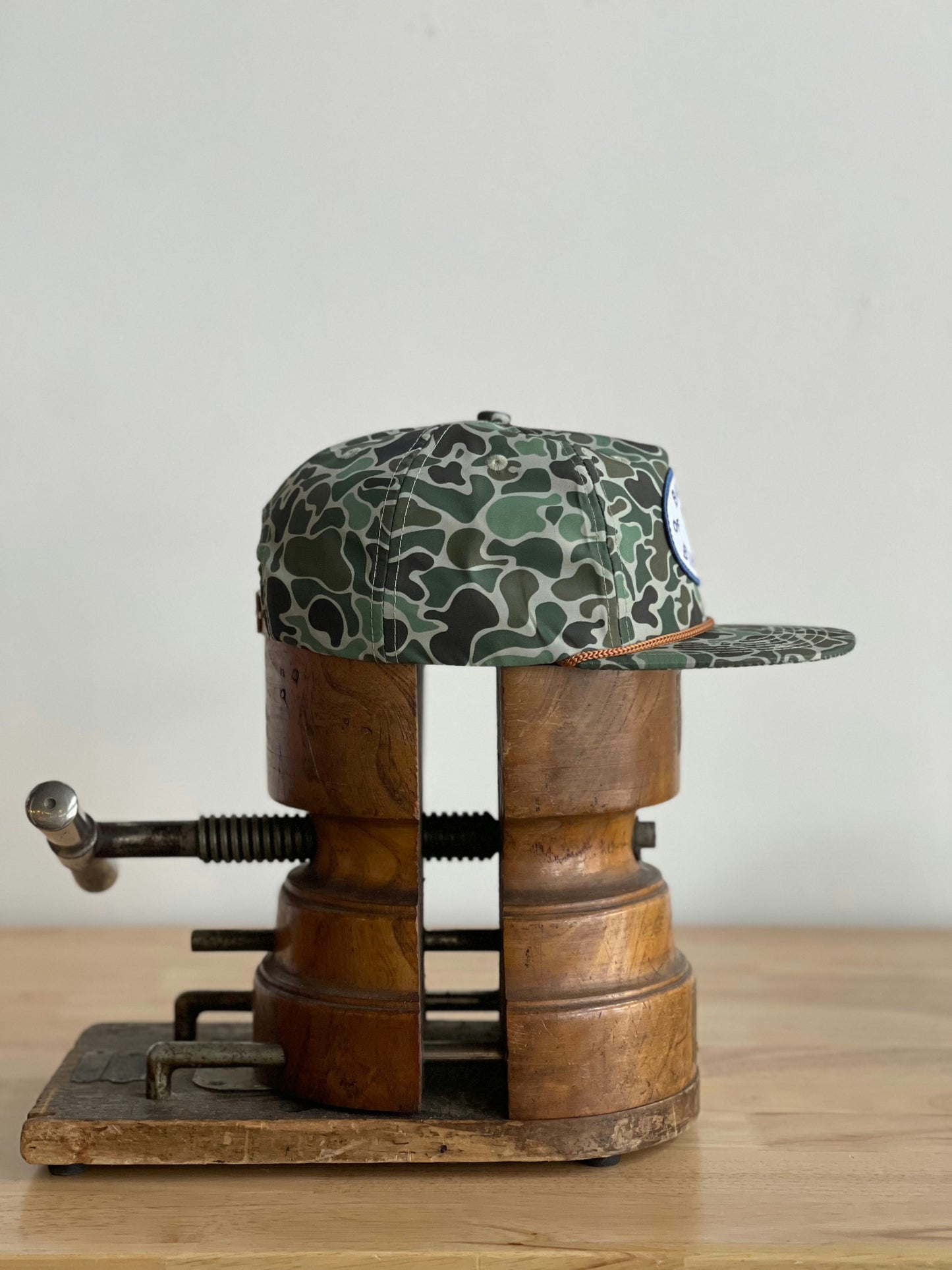 The Shop Hat | Camo Emblem by Ballad of the Bird Dog is displayed on a vintage wooden clamp atop a light wood surface, set against a plain background.