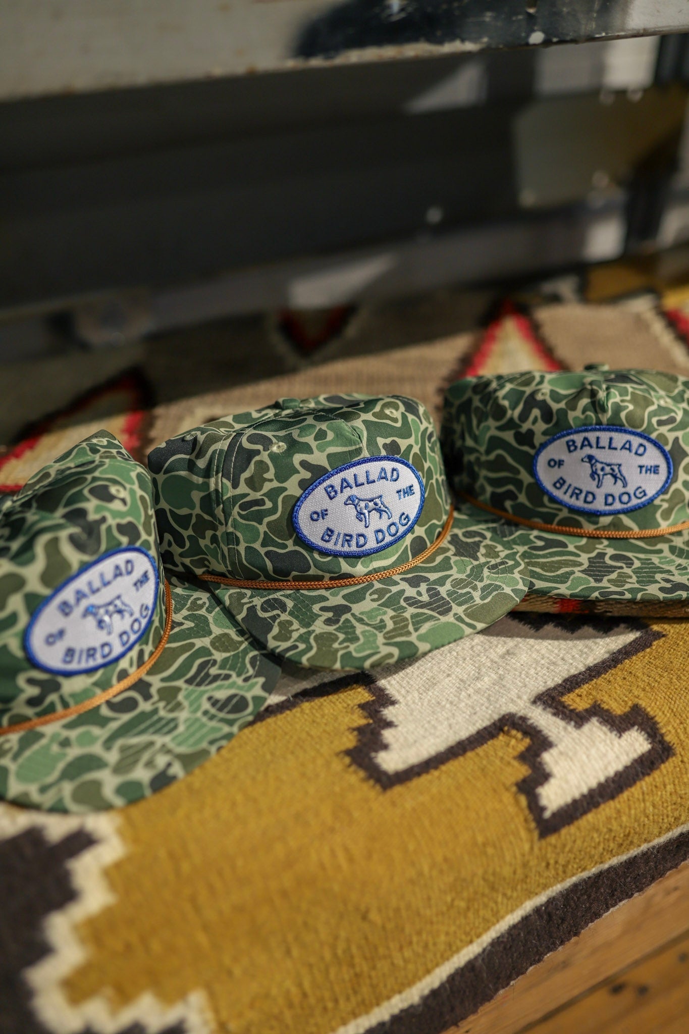 Three Shop Hat | Camo Emblem hats by Ballad of the Bird Dog, featuring blue and white patches with "Ballad of the Bird Dog," are displayed on a patterned fabric surface.