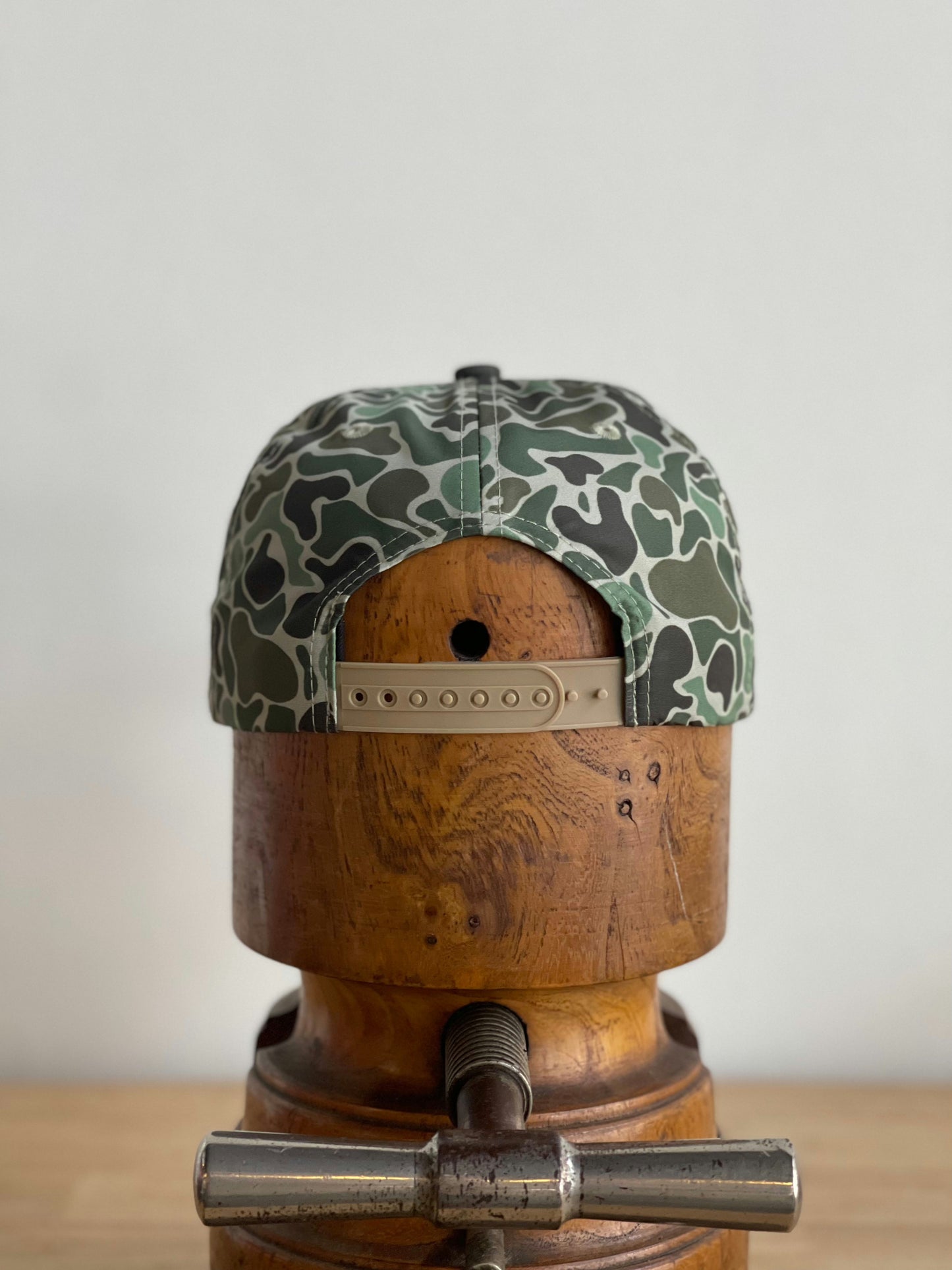 The Ballad of the Bird Dog Shop Hat | Camo Emblem is shown from the back on a wooden hat block, featuring a green camouflage snapback design.