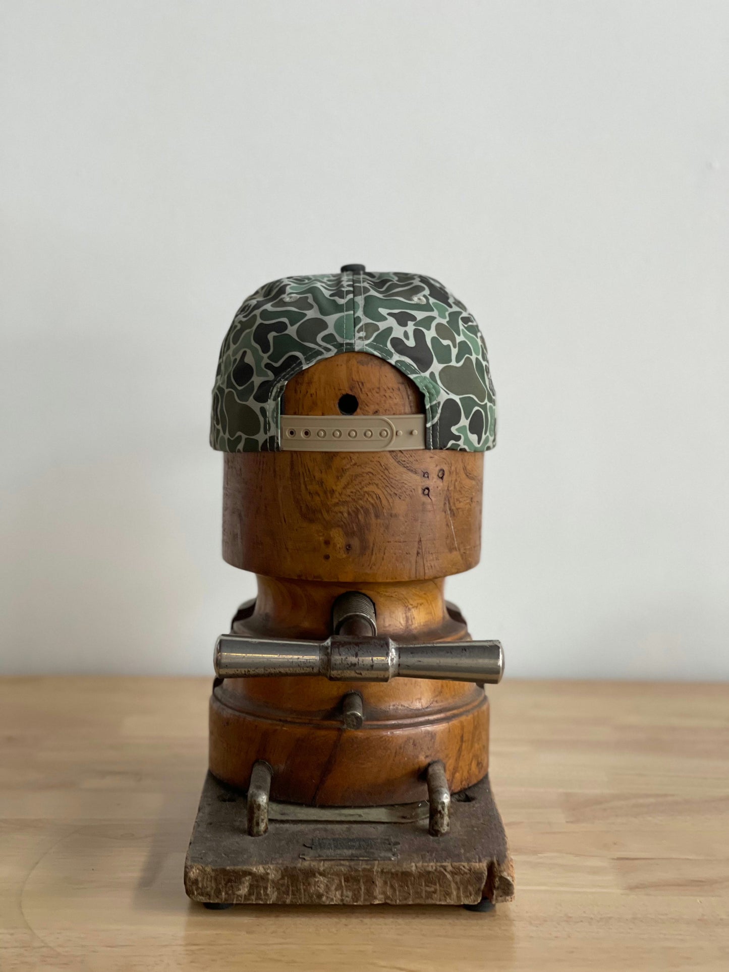 The Shop Hat | Camo Emblem by Ballad of the Bird Dog is shown backwards on a vintage wooden hat block, placed on a light wood table against a plain white background.