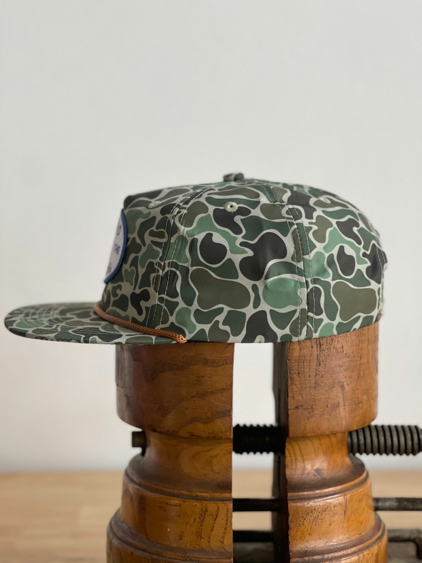 The Shop Hat | Camo Emblem by Ballad of the Bird Dog rests securely on a wood clamp, showcasing its rugged style in a practical display.