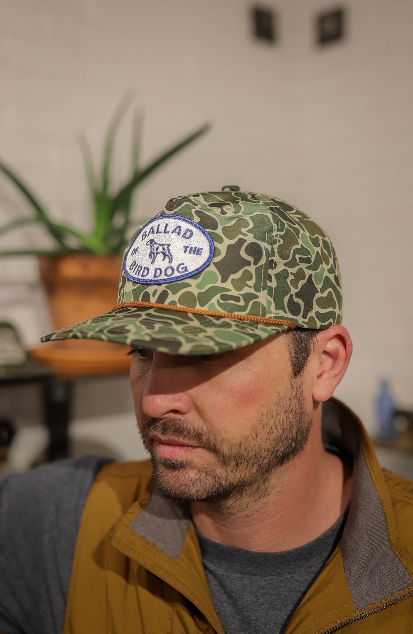 A man indoors, with a potted plant behind him, wears the Ballad of the Bird Dog Shop Hat | Camo Emblem along with a brown vest and gray shirt.