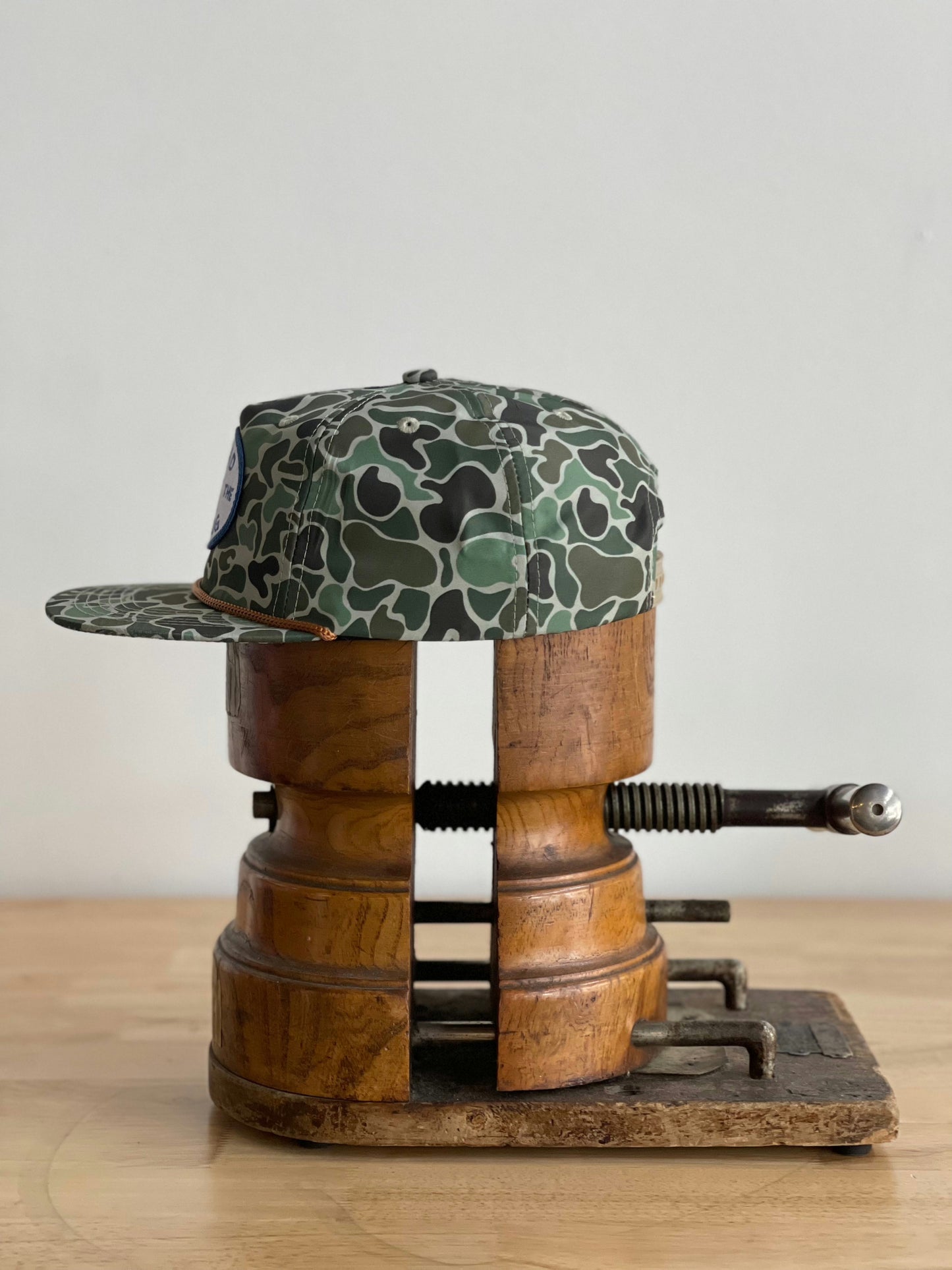 The Shop Hat | Camo Emblem by Ballad of the Bird Dog is shown in a wooden hat stretcher, set on a light wood surface with a plain background.