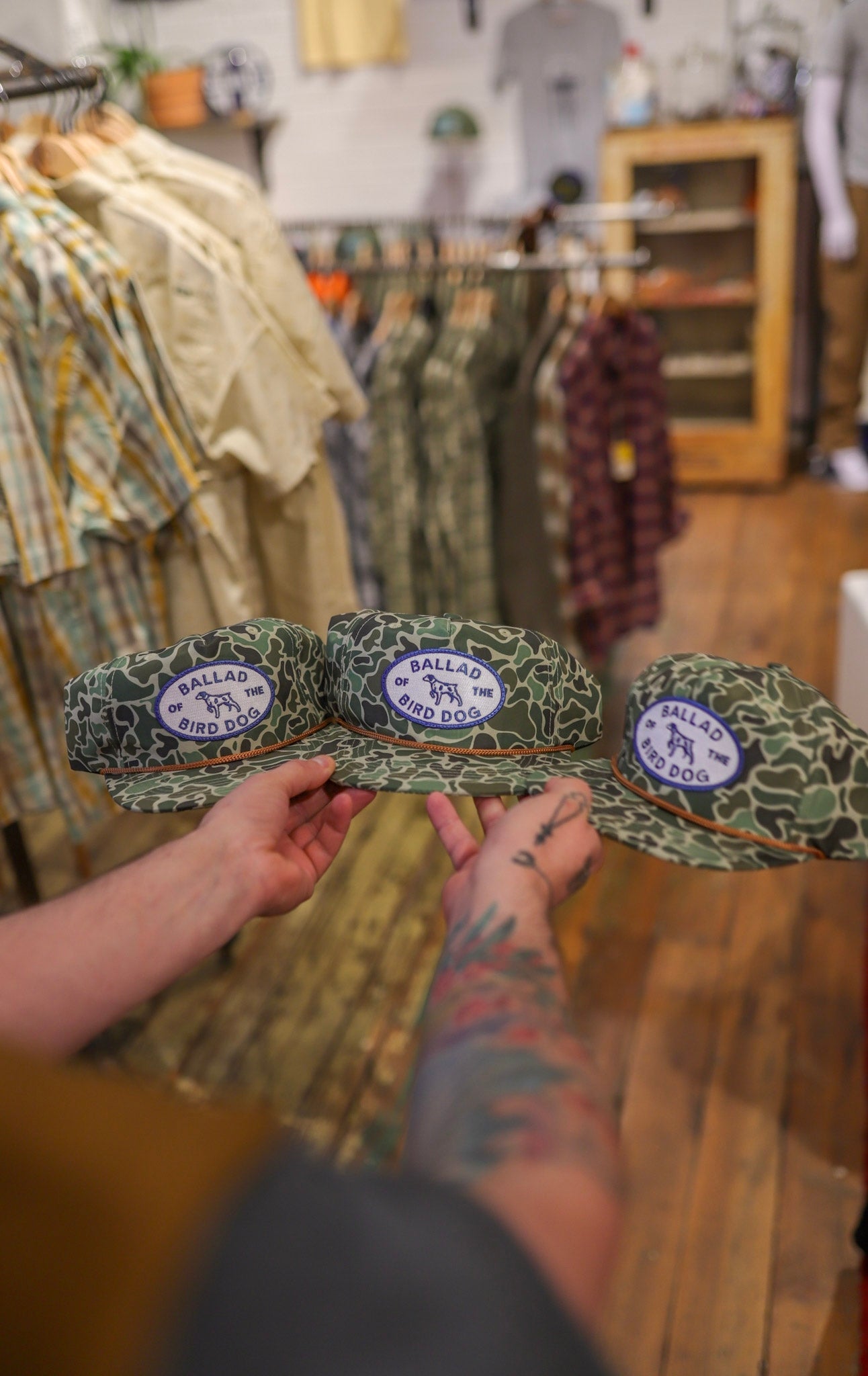A person with tattooed arms holds three green camouflage hats, including the Shop Hat | Camo Emblem by Ballad of the Bird Dog, in a clothing store.