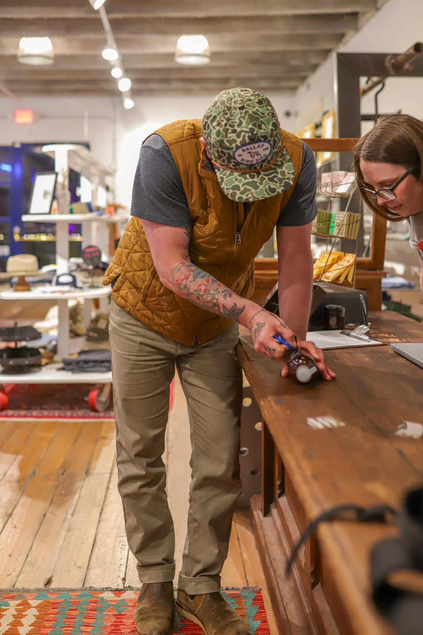 A man in a Shop Hat | Camo Emblem by Ballad of the Bird Dog and a brown vest signs a bottle at a wooden counter while a woman watches in the store.
