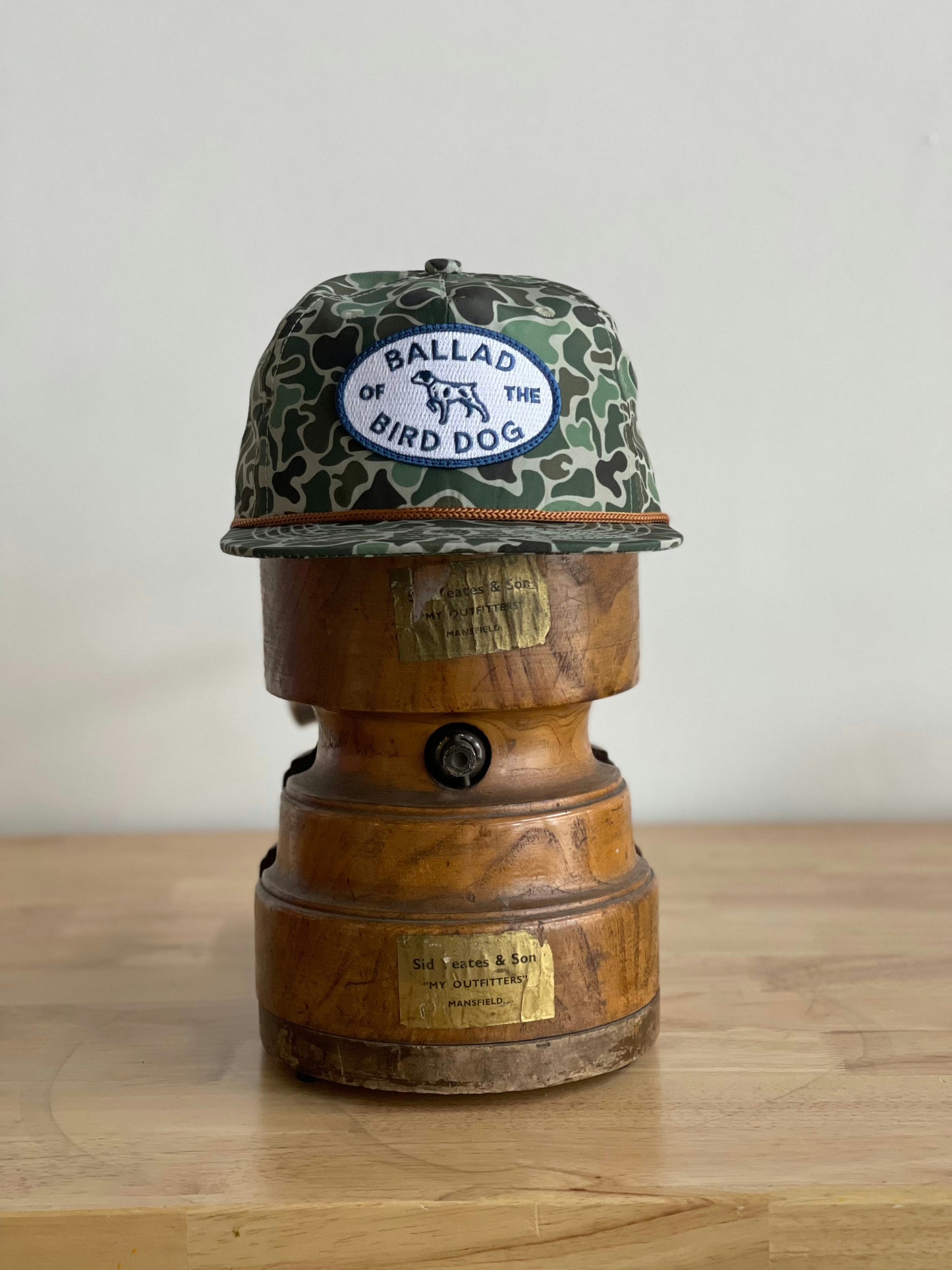 The Shop Hat | Camo Emblem by Ballad of the Bird Dog features a signature patch and sits on a vintage wood stand, creating a rustic and distinctive display for this unique bird dog hat.