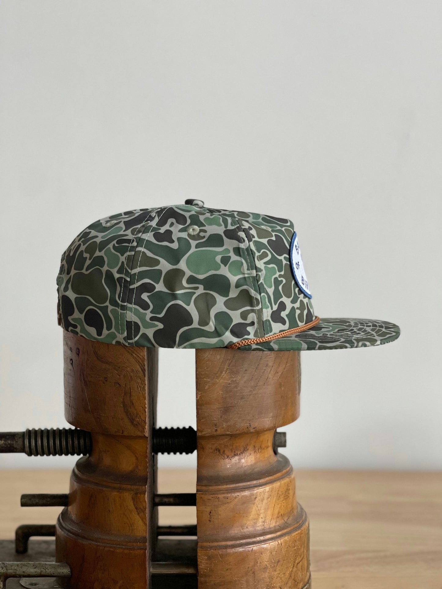 The Shop Hat | Camo Emblem by Ballad of the Bird Dog is shown in side profile on two wooden cylindrical stands against a plain background.