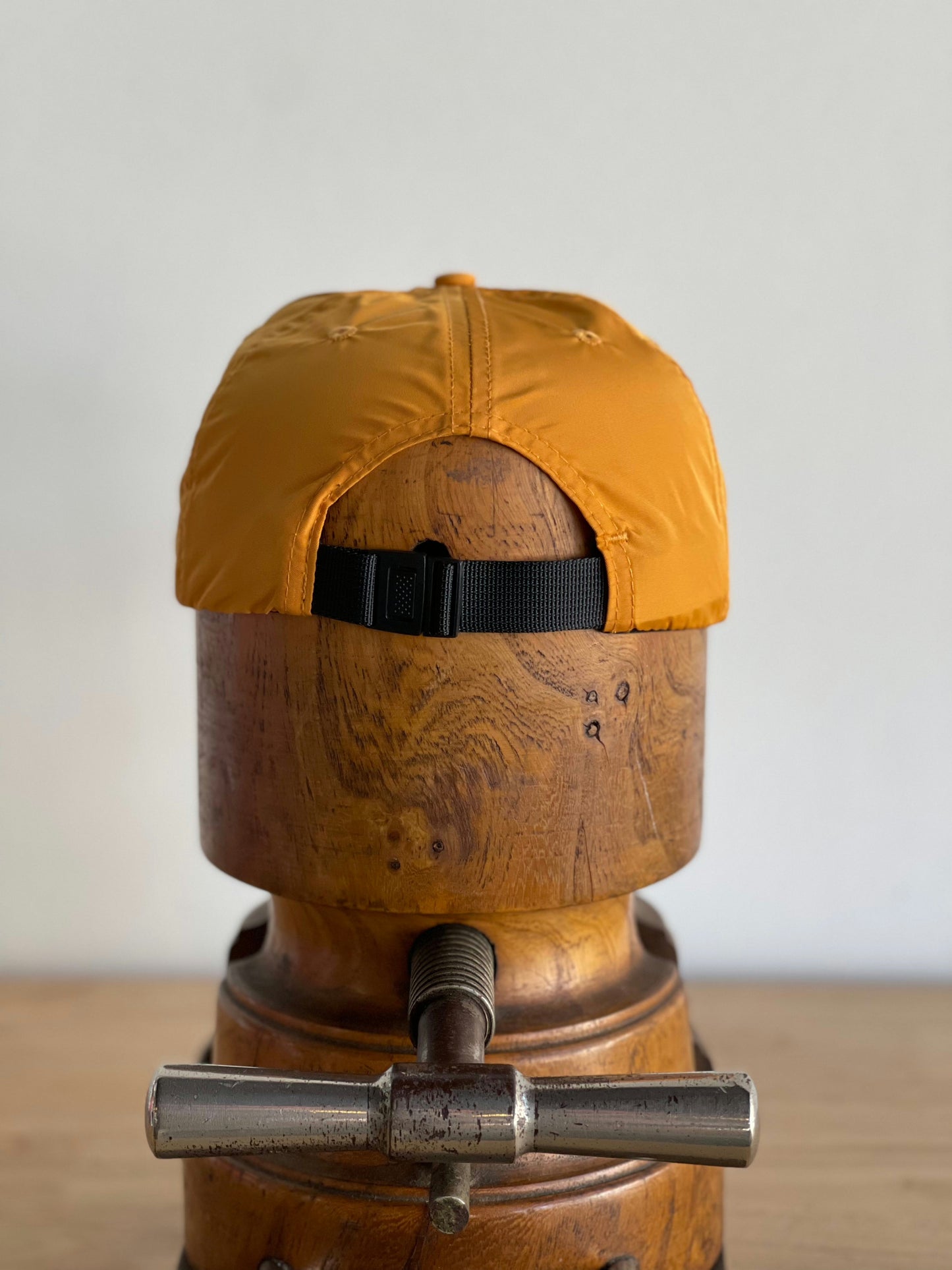 A yellow Ballad of the Bird Dog Shop Hat | Circle Clubhouse Tradition, featuring a black buckle strap, is shown from the back on a wooden stand against a plain background.