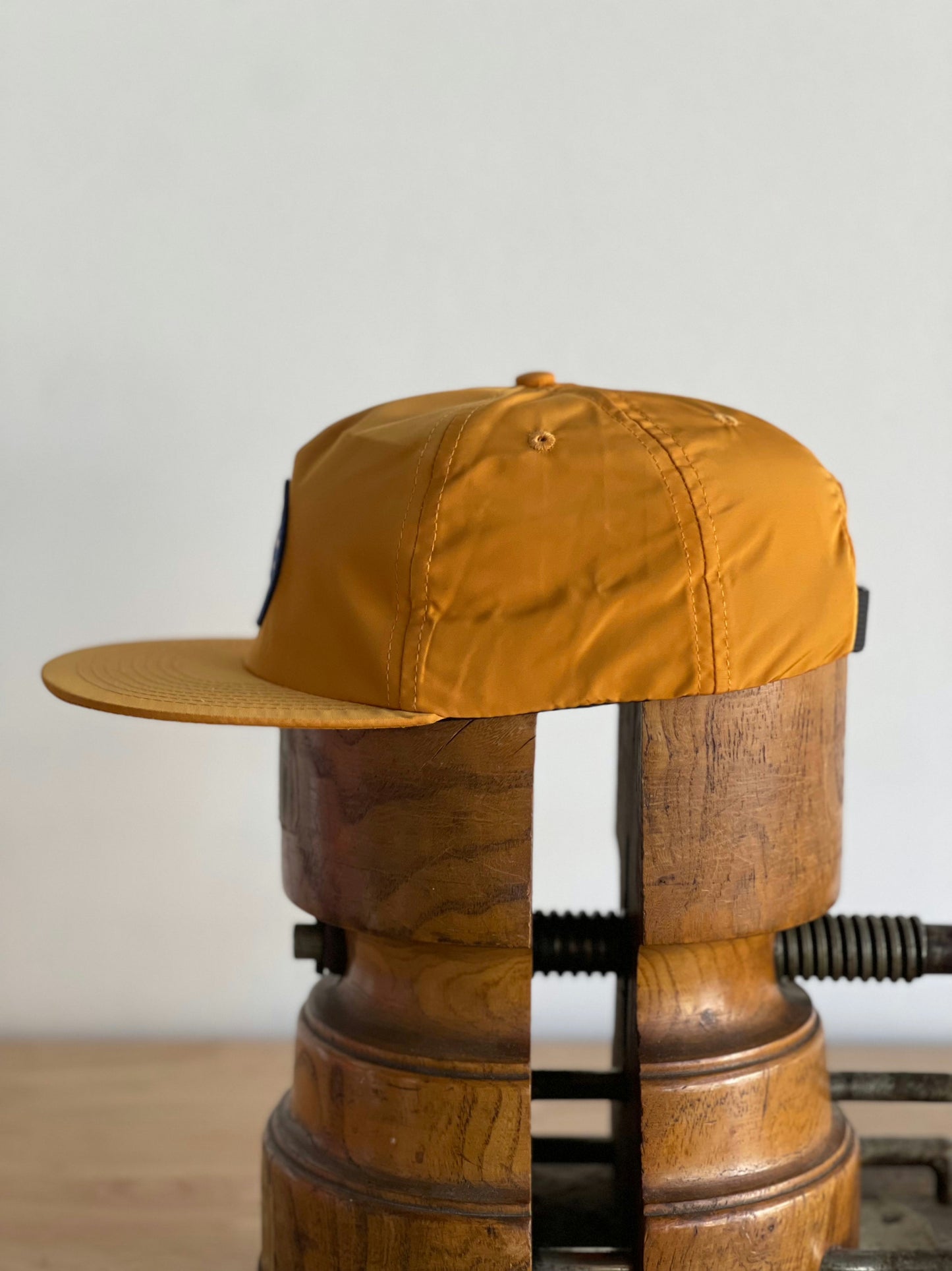 The Ballad of the Bird Dog Shop Hat, Circle Clubhouse Tradition edition, in mustard yellow with a circle logo patch, is displayed sideways on a wooden clamp against a plain background.