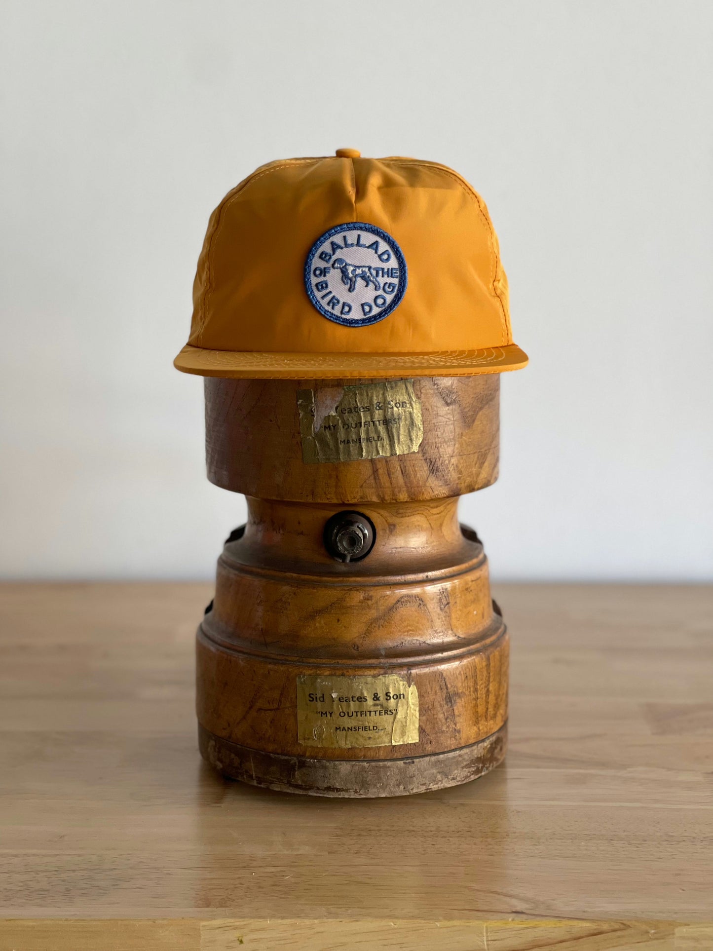 The Ballad of the Bird Dog Shop Hat | Circle Clubhouse Tradition, featuring a "BALLARD BIRD DOG OUTFITTER" patch, sits atop a vintage wooden object with metal plaques on a wooden surface.