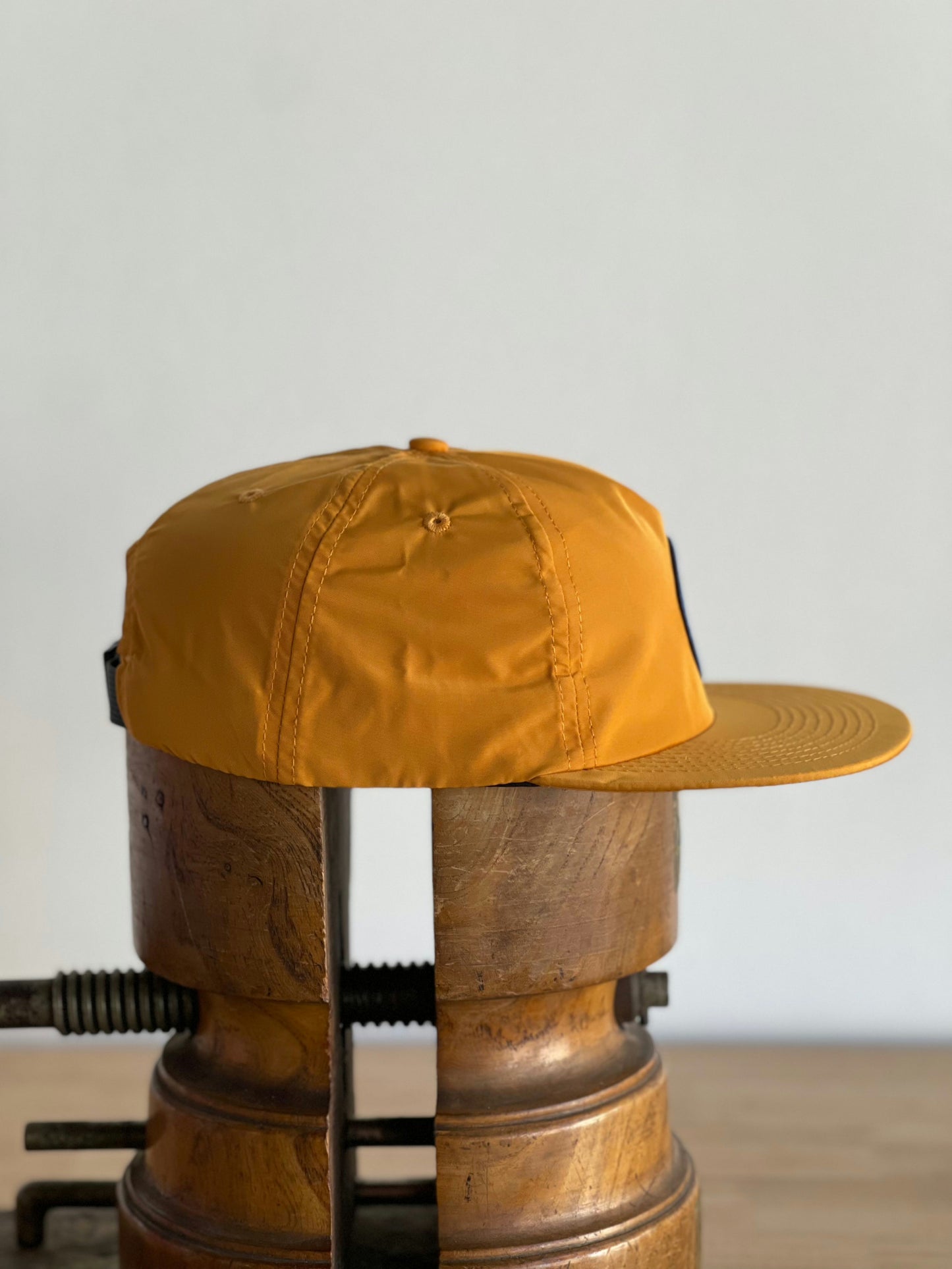 The Shop Hat | Circle Clubhouse Tradition by Ballad of the Bird Dog, a yellow patch hat, sits on two clamped wooden cylinders against a plain background.