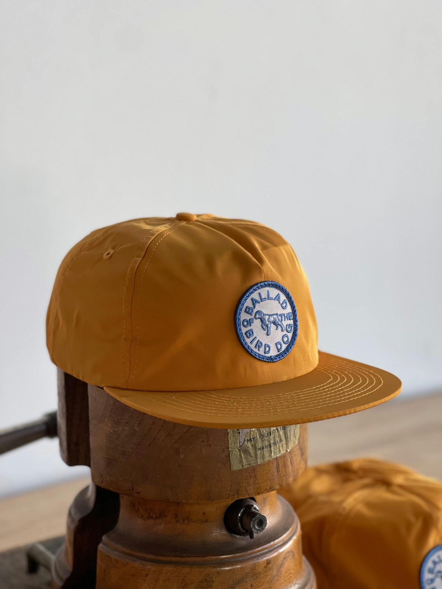 A mustard yellow Shop Hat by Ballad of the Bird Dog, featuring a “Circle Clubhouse Tradition” patch on the front, displayed on a wooden head form—classic style from Ballad of the Bird Dog.