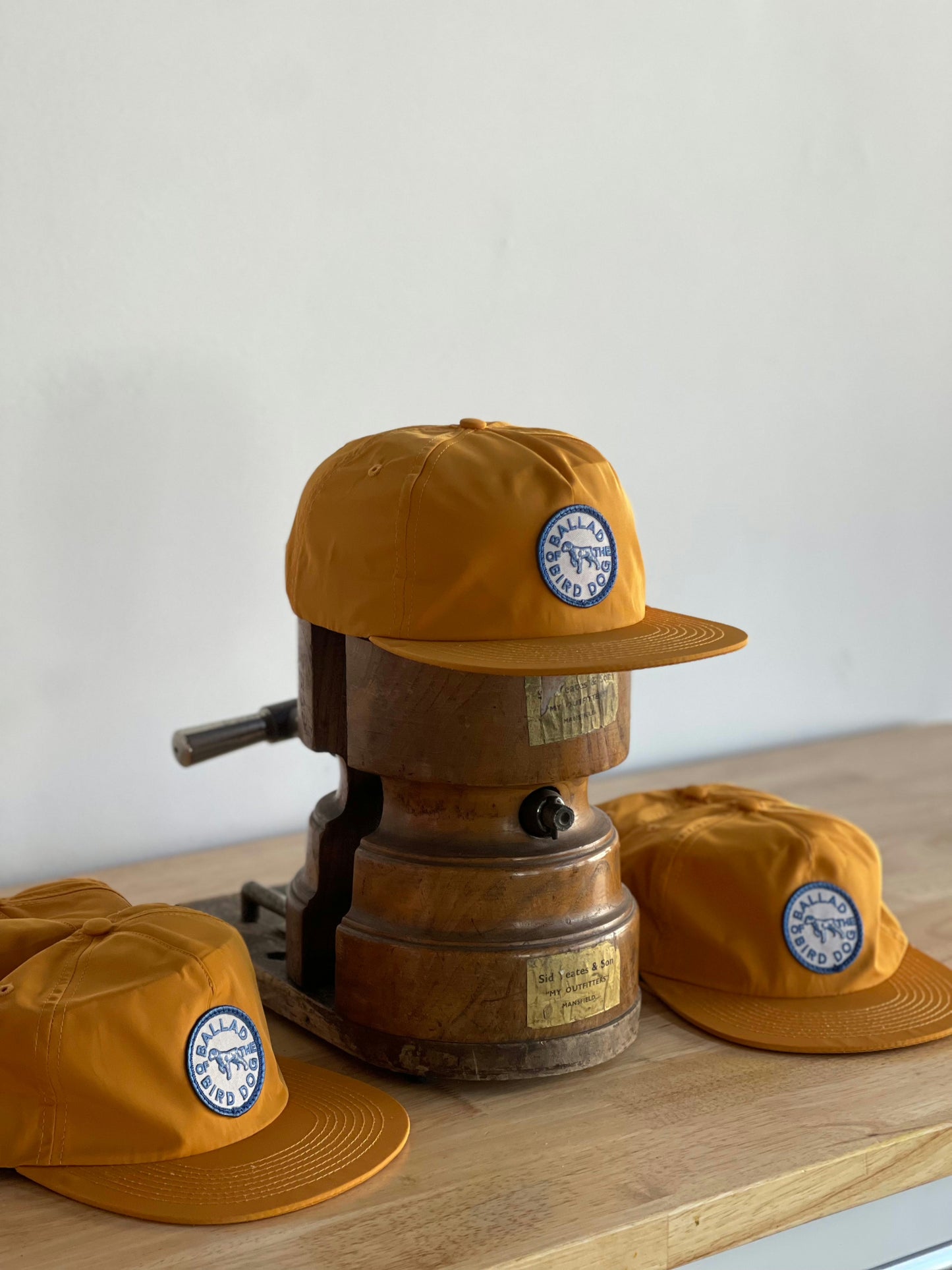 Three Ballad of the Bird Dog Shop Hat | Circle Clubhouse Tradition caps are shown, with one mustard yellow hat featuring a circle logo patch propped on a vintage metal press atop a wooden surface.