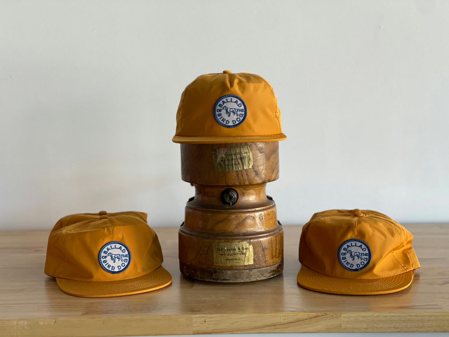 Three orange Ballad of the Bird Dog Shop Hats, each featuring a white and blue Circle Clubhouse Tradition patch, are displayed on and around two stacked antique brass weights on a wooden surface.